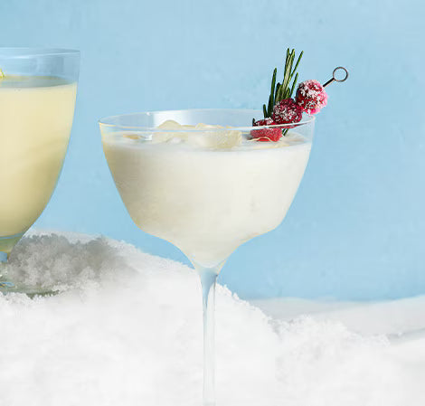 A Frozen Snow Day Colada 6 Pack cocktail, creamy white and all natural, is served in a coupe with sugared cranberries and rosemary. It sits on a snowy blue background, with another yellow party drink partially visible nearby.